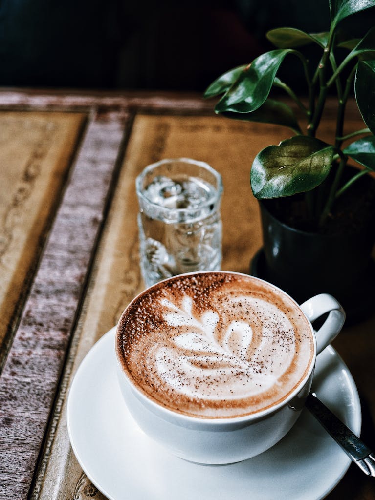 A delicious cappuccino with latte art on a rustic wooden table, accompanied by a leafy plant and water glass.