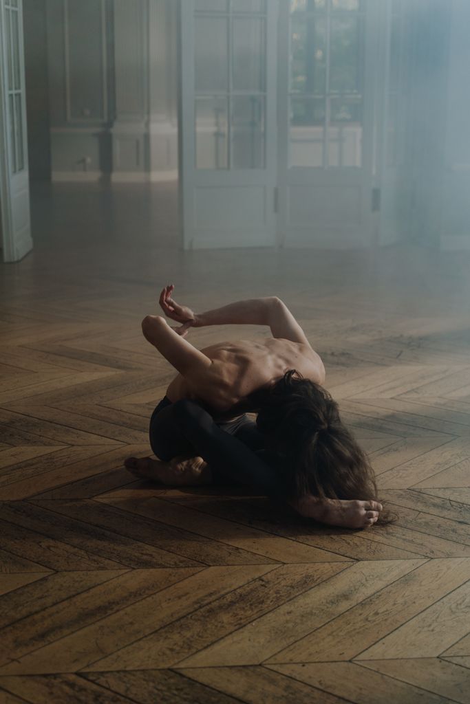 A shirtless male dancer performing an acrobatic pose indoors, showcasing flexibility and grace.