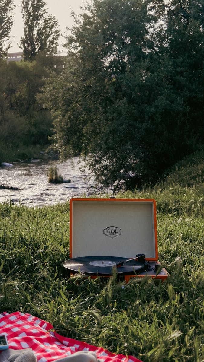 Retro record player set on a picnic blanket by a peaceful riverside, surrounded by lush greenery.